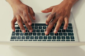 Close-up of silver latop standing on white desk, dark-skinned man working on it, young black hands, typing text into keyboard, tapping fingers on computer