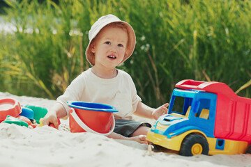 Boy on the beach near the water pours sand into a bucket with a shovel, surrounded by toys, deeply focused on the activity.