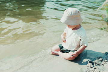 Cute boy with a hat sits on the lake's edge in wet clothes, splashing water and throwing wet sand, enjoying his time by the water.