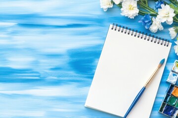 Open sketchbook rests on a blue watercolor backdrop with brush, paints, and white and blue flowers suggesting creative art and artistic endeavor