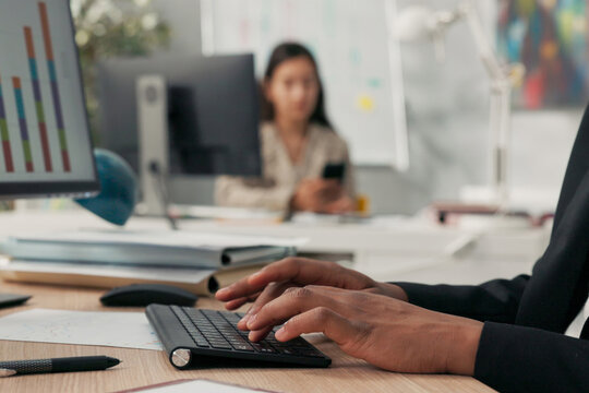 Close-up of a black keyboard from a computer standing on a wooden desk, a dark-skinned man with young hands is working at it, typing text on the keyboard, tapping fingers on the keys