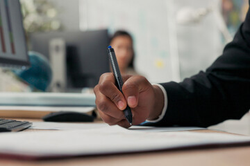 Close-up hand shot of dark-skinned man holding pen, signing documents, filling out application, cuffs of black suit, office scene, boss, manager, businessman gives consent, makes initial