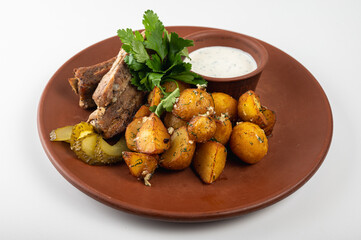 Delicious grilled pork ribs with golden roasted potatoes, pickles, and fresh parsley on a rustic clay plate. Served with creamy white sauce. Perfect homemade meal	
