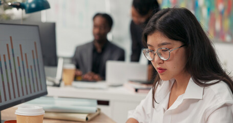Fototapeta premium An attractive girl wearing glasses works at a desk in front of a computer, fills out documents, signs checks, in the background co-workers are browsing the project on the computer, talking, discussing