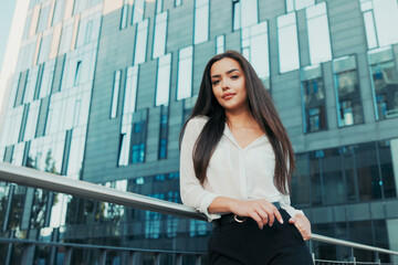 Beautiful attractive elegantly dressed in white shirt woman stands at railing next to glass building of corporation where she works, businesswoman looks ahead, hair disheveled, smiling at camera