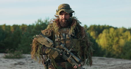 A determined soldier in camouflage uniform and helmet unholsters his rifle, standing at attention with a firm grip on his weapon, raising his eyebrows in a defiant and confident gaze.