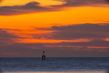 A striking seascape capturing the vivid hues of a sunset sky blending orange, red, and purple over the calm ocean. A navigation tower stands against the horizon, adding depth and dramatic scene.  .