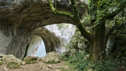 Scenic view of a natural stone arch cave surrounded by rugged rock formations and lush greenery. 