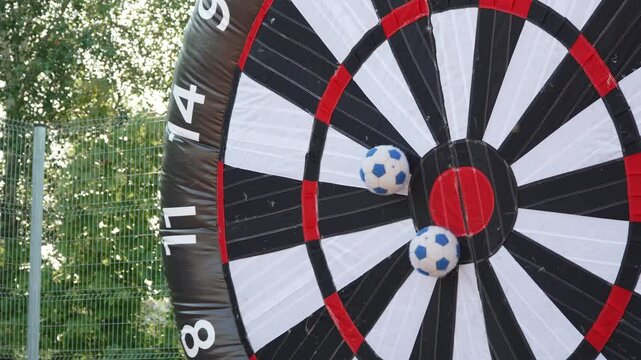 Playing with soccer balls in giant darts board during outdoor corporate party. Fun team building game of hitting center of inflatable attraction on sunny park. Ball sticks near center of target.