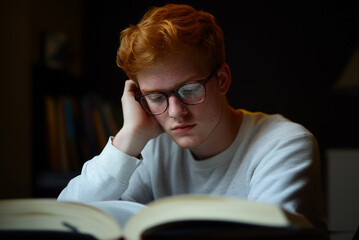 young man adjusting his glasses while reading a thick study book, lost in though