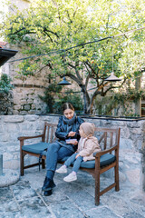 Mom feeds porridge from a spoon to a little girl sitting on a wooden bench in the courtyard of an old stone house
