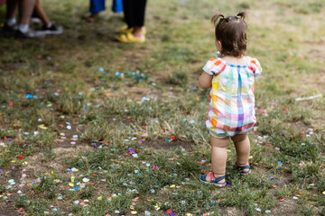 back of a cute toddler girl standing to the side of a group of people at a summer picnic. She is wearing a brightly colored checkered romper and colorful sandals with her hair in pigtails