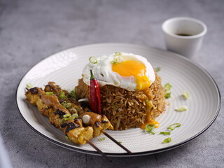 Nasi goreng with fried egg, chicken satay skewers, and soy dipping sauce, served on a plate with garnish.