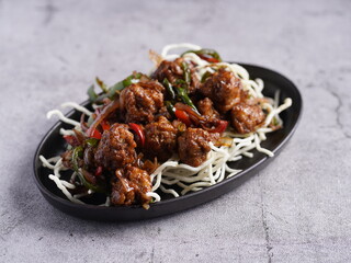 Spicy stir-fried chicken with vegetables served on crispy fried noodles in a black oval plate, styled in Asian fusion restaurant presentation