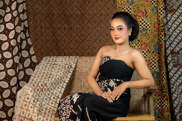 Fototapeta premium Indonesian woman in a black batik dress, sitting elegantly on a wooden bench. She exudes grace with a traditional hairstyle, vintage makeup, and cultural charm in a classic setting.