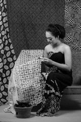Fototapeta premium An Indonesian woman in traditional batik attire creating batik using a canting. She sits elegantly on a wooden bench, surrounded by handmade fabrics, showcasing cultural heritage.