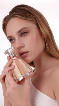 Beautiful woman in white top, blond hair and neat make-up poses in front of camera with perfume bottle in hand. Beauty face.