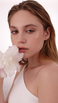 Beautiful woman in white top, blonde hair and neat makeup poses in front of camera with lily flower in hand. Beauty face.