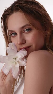 Beautiful woman in white top, blonde hair and neat makeup poses in front of camera with lily flower in hand. Beauty face.
