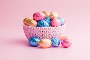 Chocolate easter eggs wrapped in multi colored foil in bowl on pink background.