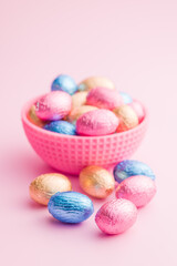 Chocolate easter eggs wrapped in multi colored foil in bowl on pink background.
