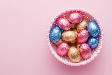 Chocolate easter eggs wrapped in multi colored foil in bowl on pink background. Top view.