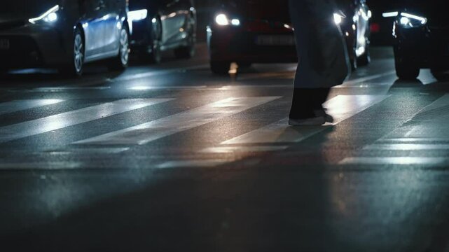 Low angle close-up female pedestrian wearing jeans and white sneakers walking street, sport shoes steps. Woman run crosswalk backlit with bright cars headlights. Evening darkness asphalt zebra