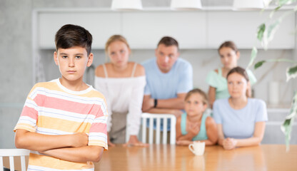 Offended pre-teen boy standing with arms crossed over chest at kitchen on background of his big family