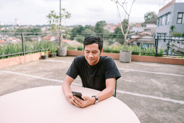 Fototapeta premium Young man using cellphone at outdoor rooftop with cityscape view