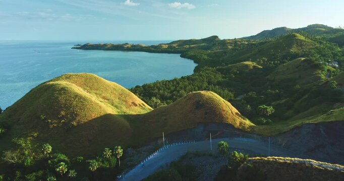 Lush green hills meet turquoise waters in a breathtaking aerial view of Kelor island, showcasing the pristine beauty of Komodo national park, Indonesia, with a winding road and hints of vegetation
