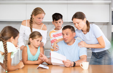 Large family is standing next to confused man sitting near kitchen table. Man reads letter and is upset by bad news from collector, unexpected test results, increase in loan rate