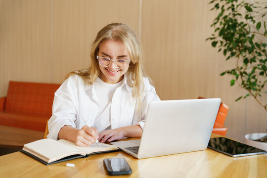 Young woman writing in notebook while working on laptop in bright, modern workspace during daytime
