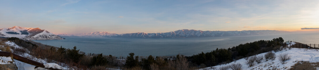 Panoramic photo of snow-covered peaks of the Sevan mountain range, as seen from Sevanavank, rise above Lake Sevan in Armenia. A breathtaking winter sunrise landscape with mist and soft sunlight