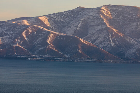 Snow-covered peaks of the Sevan mountain range, as seen from Sevanavank, rise above Lake Sevan in Armenia. A breathtaking winter sunrise landscape with mist and soft sunlight