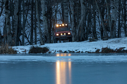 A red 4x4 off-road SUV parked near the frozen Gosh Lake in Dilijan National Park, Tavush, Armenia. The headlights reflect on the icy surface, creating a rugged winter adventure scene