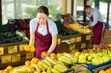 With practiced hand, woman supermarket worker arrange oranges in neat pyramid by window. Female seller of vegetable department of store is replenishing showcase with oranges, puts ones on showcase