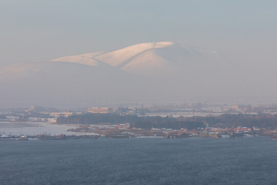 Snow-covered peaks of the Geghama mountain range,  rise above Lake Sevan and city Sevan in Armenia. A breathtaking winter sunrise landscape with mist and soft sunlight