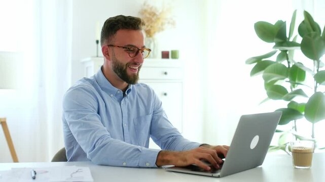Achievement goal. Cheerful successful businessman with glasses in a blue shirt, raising his fists while looking at his laptop screen, celebrating a success in a bright modern office, smiling happily