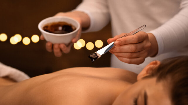 Unrecognizable massage therapist applying exfoliating body mask with brush on relaxed woman back at spa, body care concept. Smiling young lady having body treatment with aroma at modern spa, closeup
