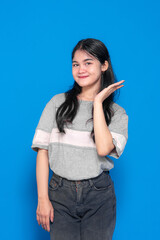 A cheerful young woman with long black hair, wearing a casual gray t-shirt and black jeans, smiling and posing playfully against a solid blue background.