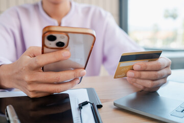 Person holding smartphone and credit card, engaging in online shopping at a home office desk with a laptop.