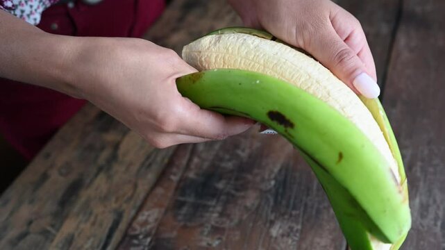 Woman peeling green plantain for patacones or tostones on wooden table