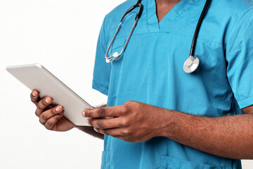 An African American man doctor stands in a hospital, holding a tablet while dressed in scrubs. He is focused on providing healthcare and ensuring efficient patient management.