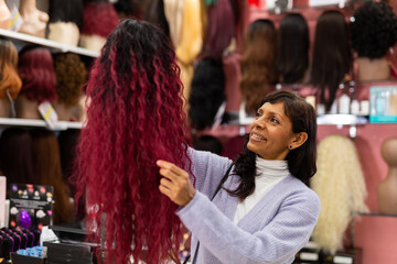 Smiling hispanic woman shopping in specialized store, choosing stylish wigs from natural hair..