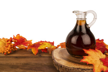 Glass jug of maple syrup placed on a rustic wooden table with vibrant autumn leaves. A cozy and warm seasonal setting, perfect for fall-themed designs, food photography, and thanksgiving visuals.