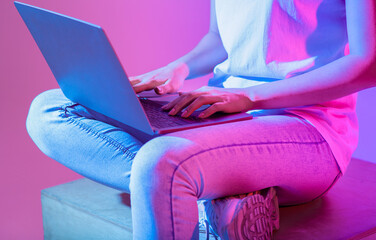 Woman sits on a block, typing on a laptop. The ambient lighting casts vibrant colors, creating a...