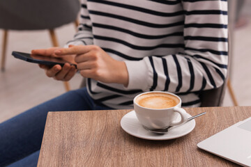 Woman using smartphone during coffee break at wooden table indoors, closeup