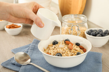 Woman pouring milk into bowl with oatmeal, blueberries and almonds at wooden table, closeup