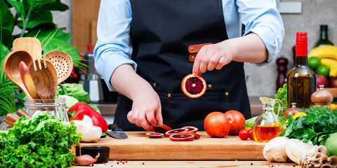 Chef holds wheel of sliced ​​blood orange in hand. Cozy kitchen with wooden table, kitchenware, vegetables, herbs and ingredients for cooking. Healthy vegan food, culinary, recipes, food blogging