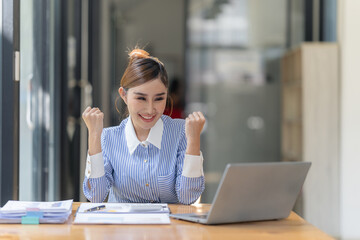 Cheerful Asian woman celebrates victory. Happy, excited, celebrating success with digital tablet and laptop in office.